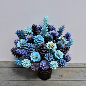 May include: A bouquet of blue and purple pine cones painted to look like flowers. The pine cones are arranged in a black vase.