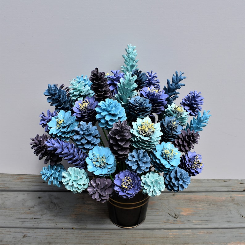 May include: A bouquet of blue and purple pine cones painted to look like flowers. The pine cones are arranged in a black vase.