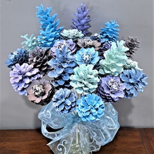 May include: Two Dozen Flowers, a bouquet of blue, purple, and green pine cones painted to look like flowers in a clear glass vase with a blue ribbon.