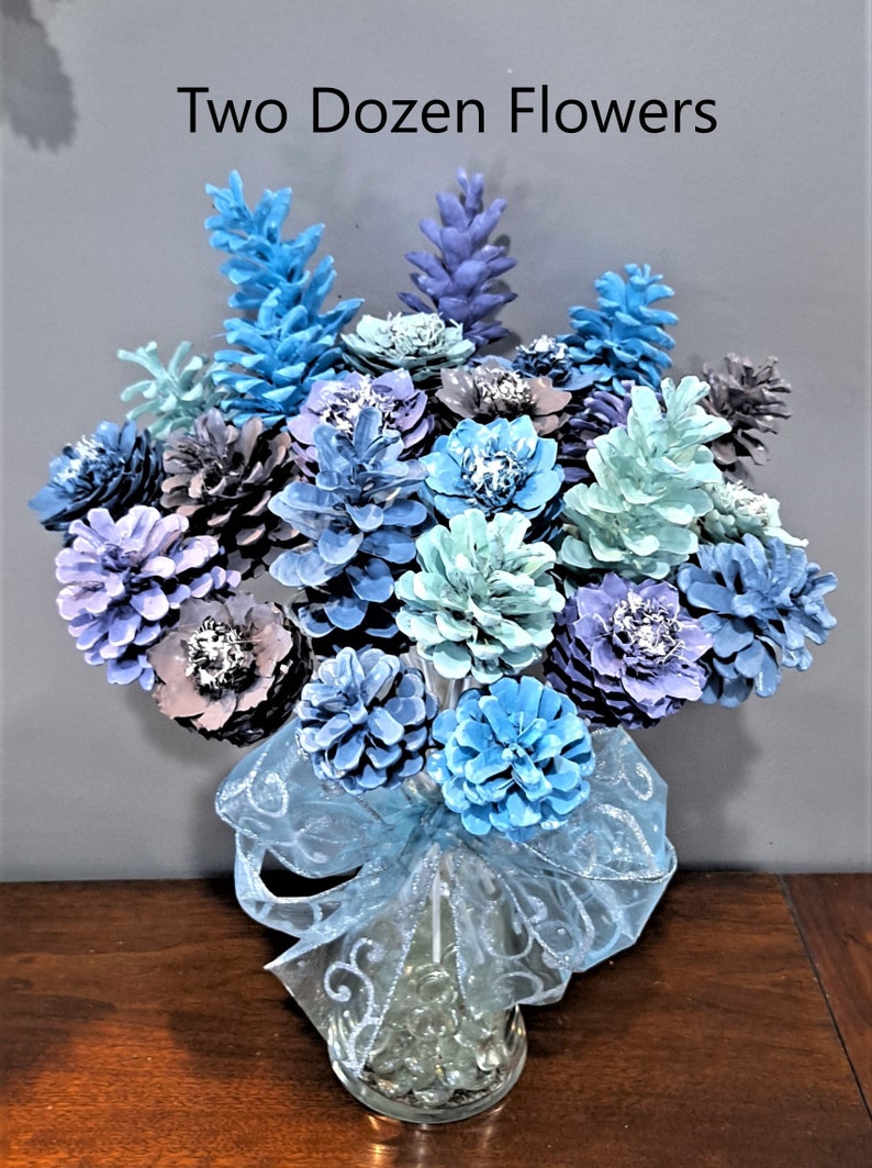 May include: Two Dozen Flowers, a bouquet of blue, purple, and green pine cones painted to look like flowers in a clear glass vase with a blue ribbon.
