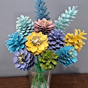 May include: A dozen painted pine cones arranged in a vase, resembling a bouquet of flowers. The pine cones are painted in various colours, including blue, green, yellow, and pink. The vase is filled with clear glass beads. The text "One Dozen Flowers (Vase is Not Included)" is displayed above the arrangement.