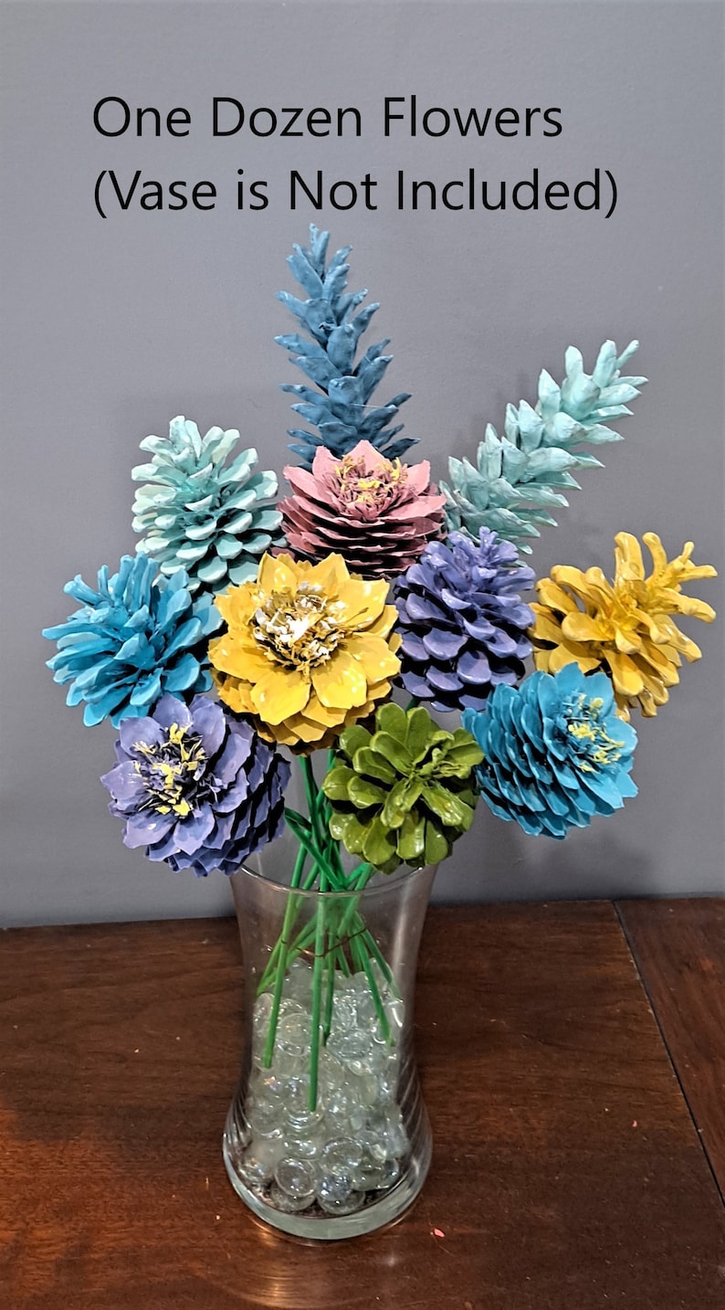 May include: A dozen painted pine cones arranged in a vase, resembling a bouquet of flowers. The pine cones are painted in various colours, including blue, green, yellow, and pink. The vase is filled with clear glass beads. The text "One Dozen Flowers (Vase is Not Included)" is displayed above the arrangement.