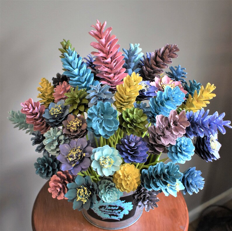 May include: A colourful bouquet of painted pine cones in a variety of shades of blue, green, pink, and yellow. The pine cones are arranged in a vase and are ready to be displayed in a home or office.