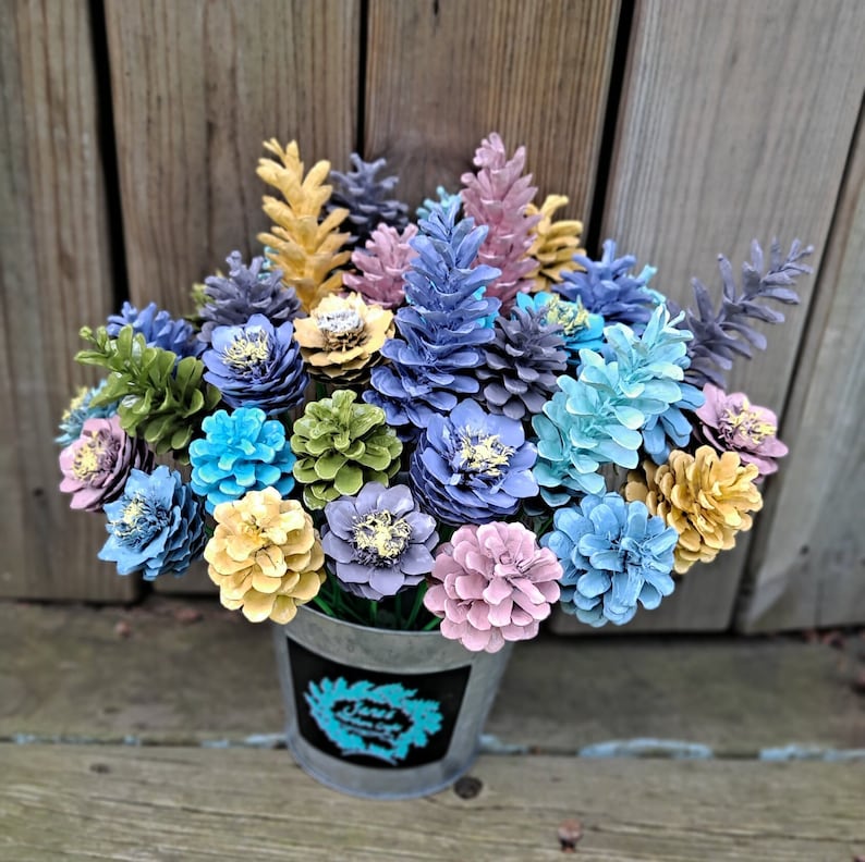 May include: A bouquet of painted pine cones in a variety of colours, including blue, green, yellow, and pink. The pine cones are arranged in a metal vase.