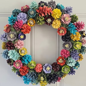 May include: A vibrant, colorful wreath composed of painted pinecones in various hues, including teal, pink, yellow, and purple. The pinecones are arranged in a circular pattern, creating a decorative floral design. The wreath is suitable for seasonal home decor.