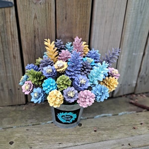 May include: A bouquet of painted pine cones arranged in a galvanised metal bucket. The pine cones are painted in various pastel colours, including blue, green, yellow, and pink. The bucket has a black label with the text "Just Breathe & Enjoy".