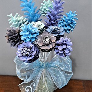 May include: A dozen pine cone flowers in shades of blue, purple, and gray arranged in a clear glass vase with blue ribbon and glass beads. The text "One Dozen Flowers Vase is Not Included" is at the top of the image.