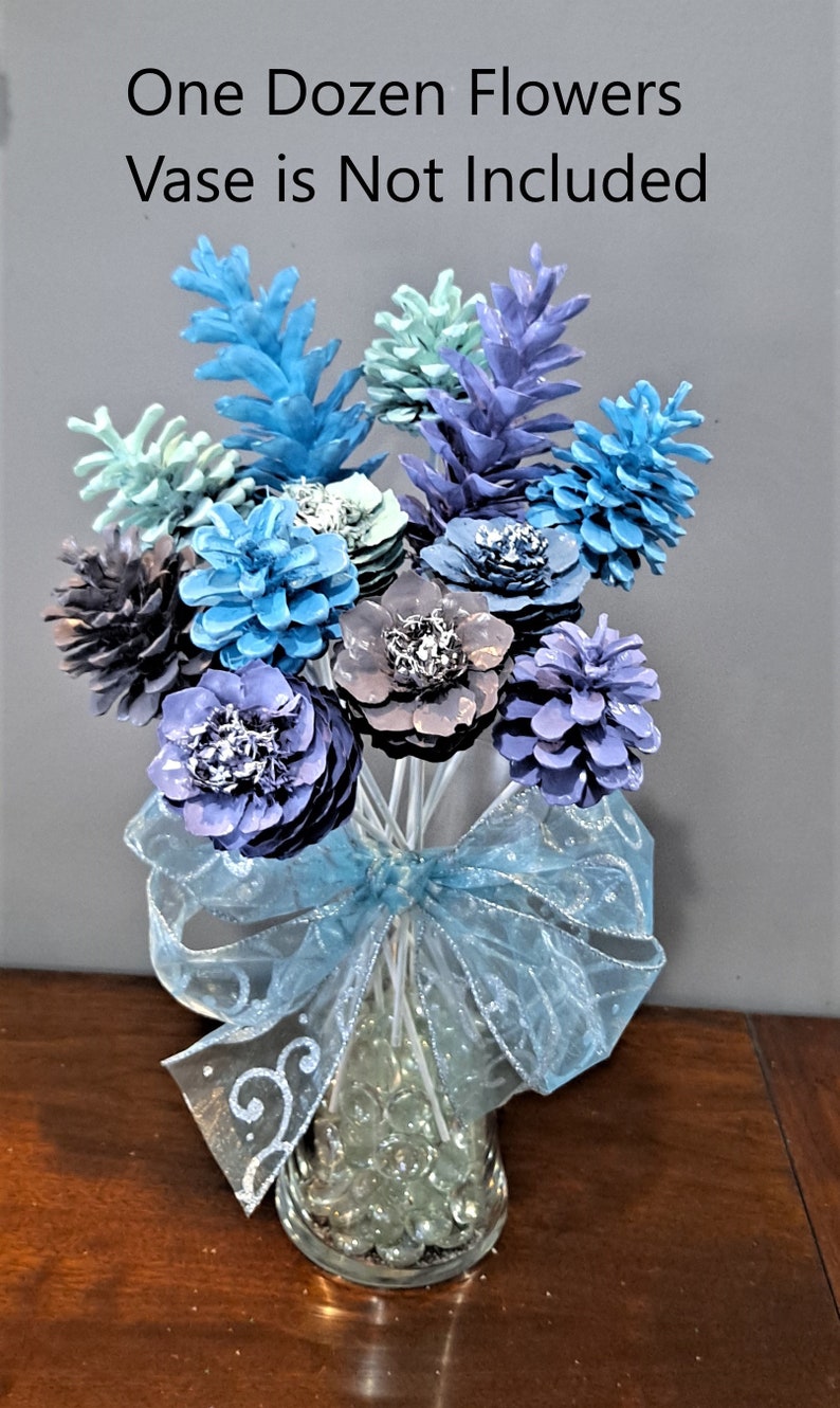 May include: A dozen pine cone flowers in shades of blue, purple, and gray arranged in a clear glass vase with blue ribbon and glass beads. The text "One Dozen Flowers Vase is Not Included" is at the top of the image.