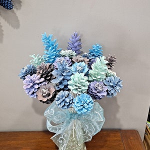 May include: A bouquet of blue and purple painted pine cones arranged in a vase with a blue and white ribbon. The pine cones are arranged to resemble flowers.