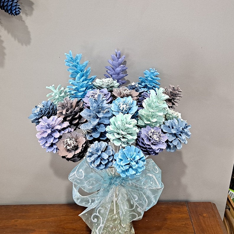 May include: A bouquet of blue and purple painted pine cones arranged in a vase with a blue and white ribbon. The pine cones are arranged to resemble flowers.