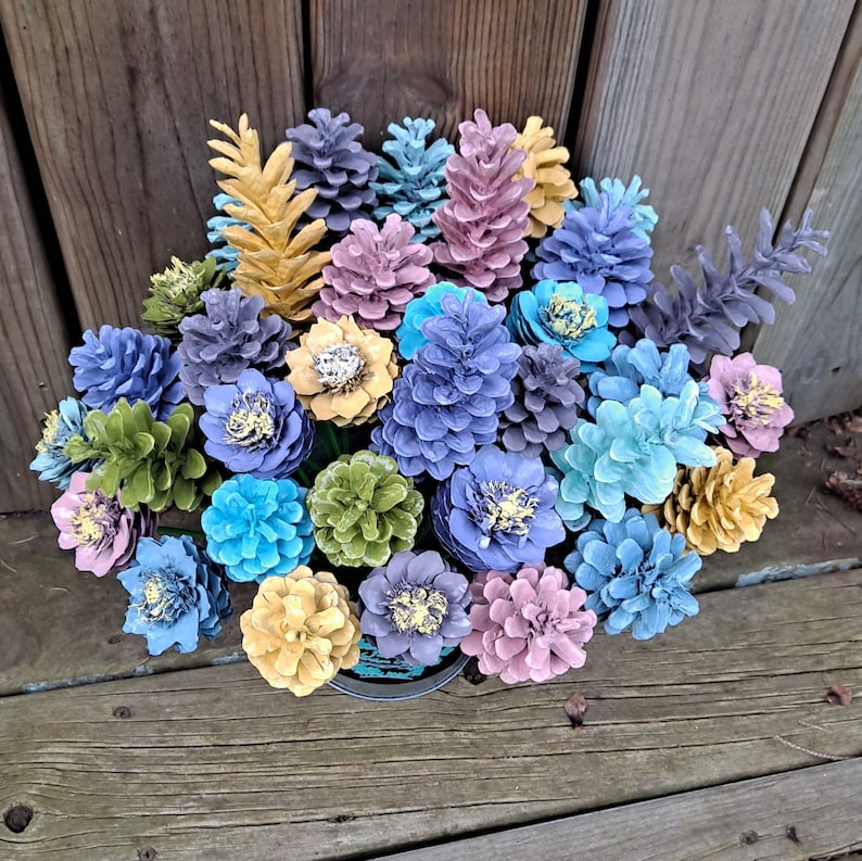 May include: A bouquet of pine cones painted in various shades of blue, green, pink, and yellow. The pine cones are arranged in a vase and create a unique and colourful floral arrangement.