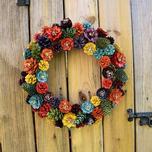 May include: A circular wreath constructed from numerous pinecones painted in a vibrant palette of red, orange, yellow, green, teal, and deep purple. Many of the pinecones feature gold-colored accents on their tips, creating a multi-colored display.