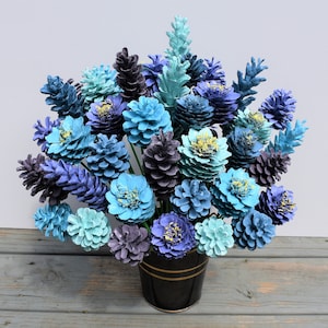 May include: A bouquet of blue and purple painted pine cones arranged in a black vase. The pine cones are painted in shades of blue, purple, and teal, and they are arranged in a vase that is black with a gold rim.
