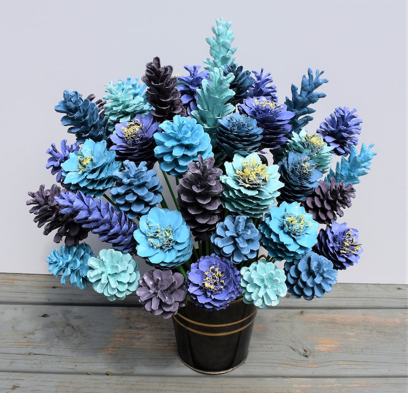 May include: A bouquet of blue and purple painted pine cones arranged in a black vase. The pine cones are painted in shades of blue, purple, and teal, and they are arranged in a vase that is black with a gold rim.
