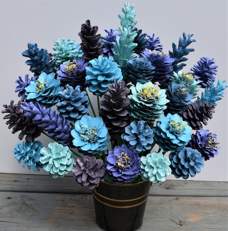 May include: A bouquet of blue and purple pine cone flowers arranged in a black vase. The flowers are made of painted pine cones and are arranged in a variety of shades of blue and purple.