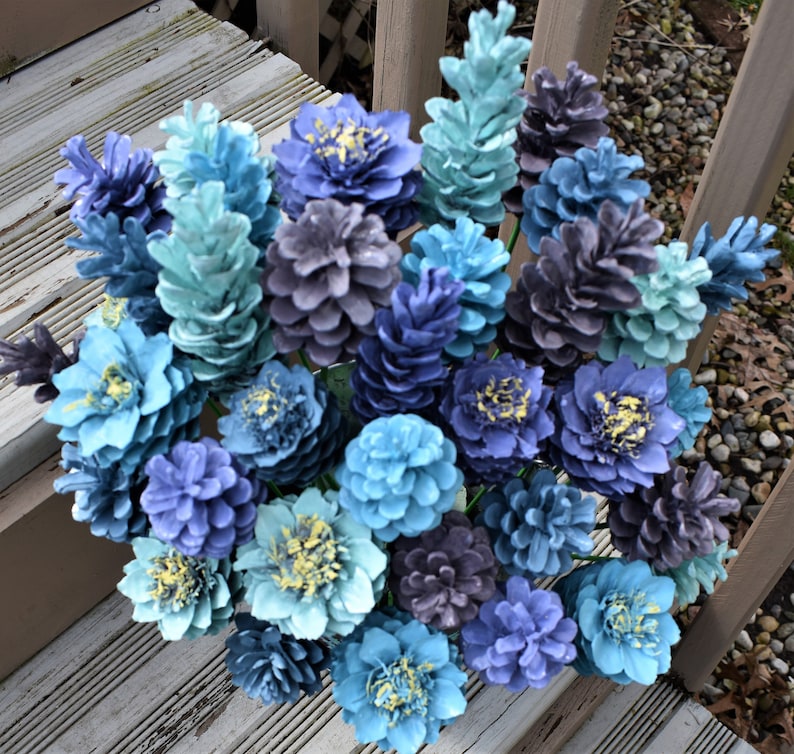 May include: A bouquet of pine cones painted in shades of blue, teal, and gray. The pine cones are arranged in a vase and look like a flower arrangement.