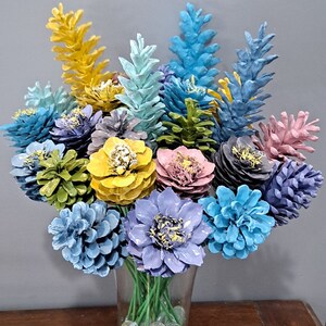 May include: A vase filled with two dozen pine cone flowers painted in various colours, including blue, yellow, green, and pink. The vase is filled with clear glass beads. The text "Two Dozen Flowers" is written above the vase.