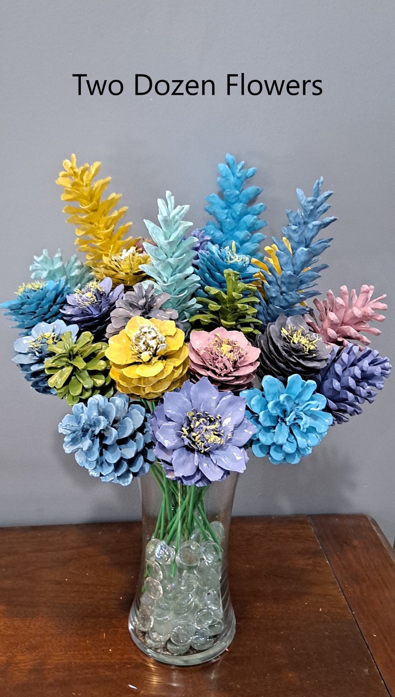 May include: A vase filled with two dozen pine cone flowers painted in various colours, including blue, yellow, green, and pink. The vase is filled with clear glass beads. The text "Two Dozen Flowers" is written above the vase.