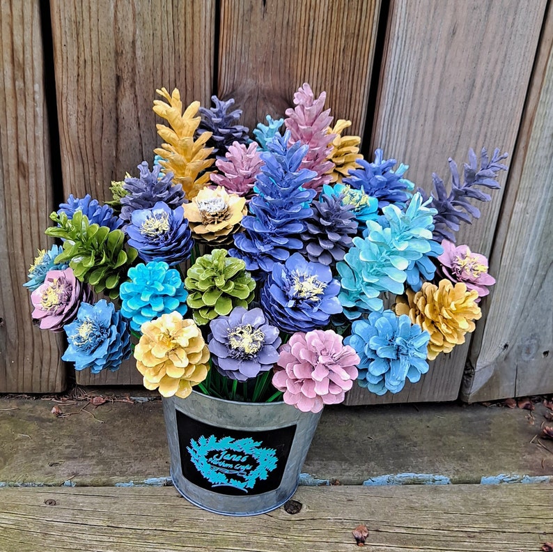 May include: A bouquet of painted pine cones arranged in a galvanised metal bucket. The pine cones are painted in shades of blue, green, yellow, and pink. The bucket has a black label with the text "Jana's Northern Crafts".