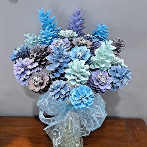 May include: A bouquet of blue and white painted pine cones arranged in a vase with a blue and white ribbon. The pine cones are arranged in a circular pattern and are all facing outwards.