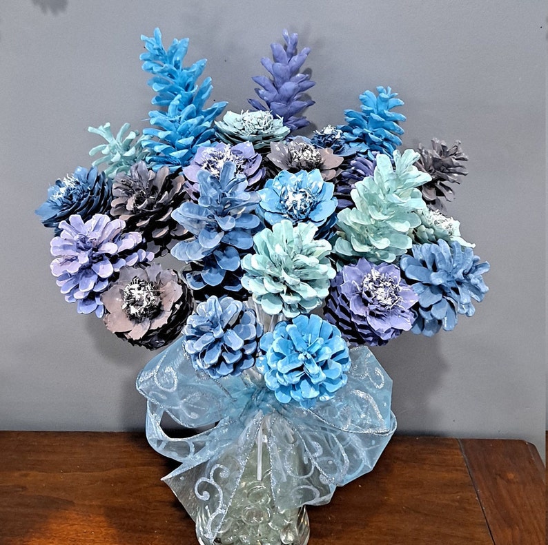 May include: A bouquet of blue and white painted pine cones arranged in a vase with a blue and white ribbon. The pine cones are arranged in a circular pattern and are all facing outwards.