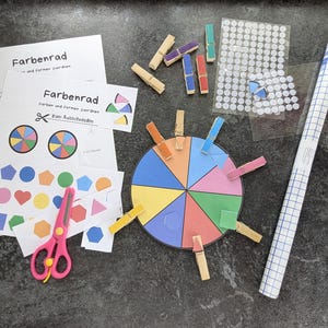 May include: An educational colour matching game featuring a colour wheel, pegs, and cut-out shapes. The game includes printed sheets with the text "Farbenrad" and a pair of pink scissors. A roll of white and blue grid paper is also included.