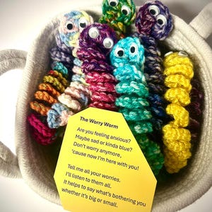Puede incluir: Una cesta llena de coloridos Worry Worms hechos a mano. Cada gusano está tejido en un color diferente y tiene ojos saltones. Una etiqueta amarilla con texto sobre los Worry Worms está dentro de la cesta.