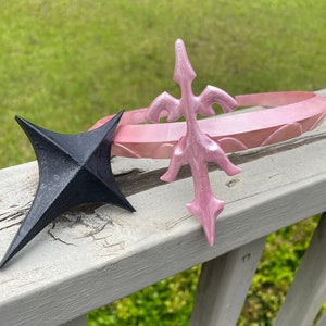 以下が含まれることがあります： 尖ったデザインのピンクのプラスチック製の王冠と、黒い星型のオブジェクトです。