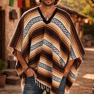 May include: A brown, tan, and white striped poncho with fringe. The poncho has a V-neck and is worn with a brown hat and blue jeans. The image is taken outdoors.