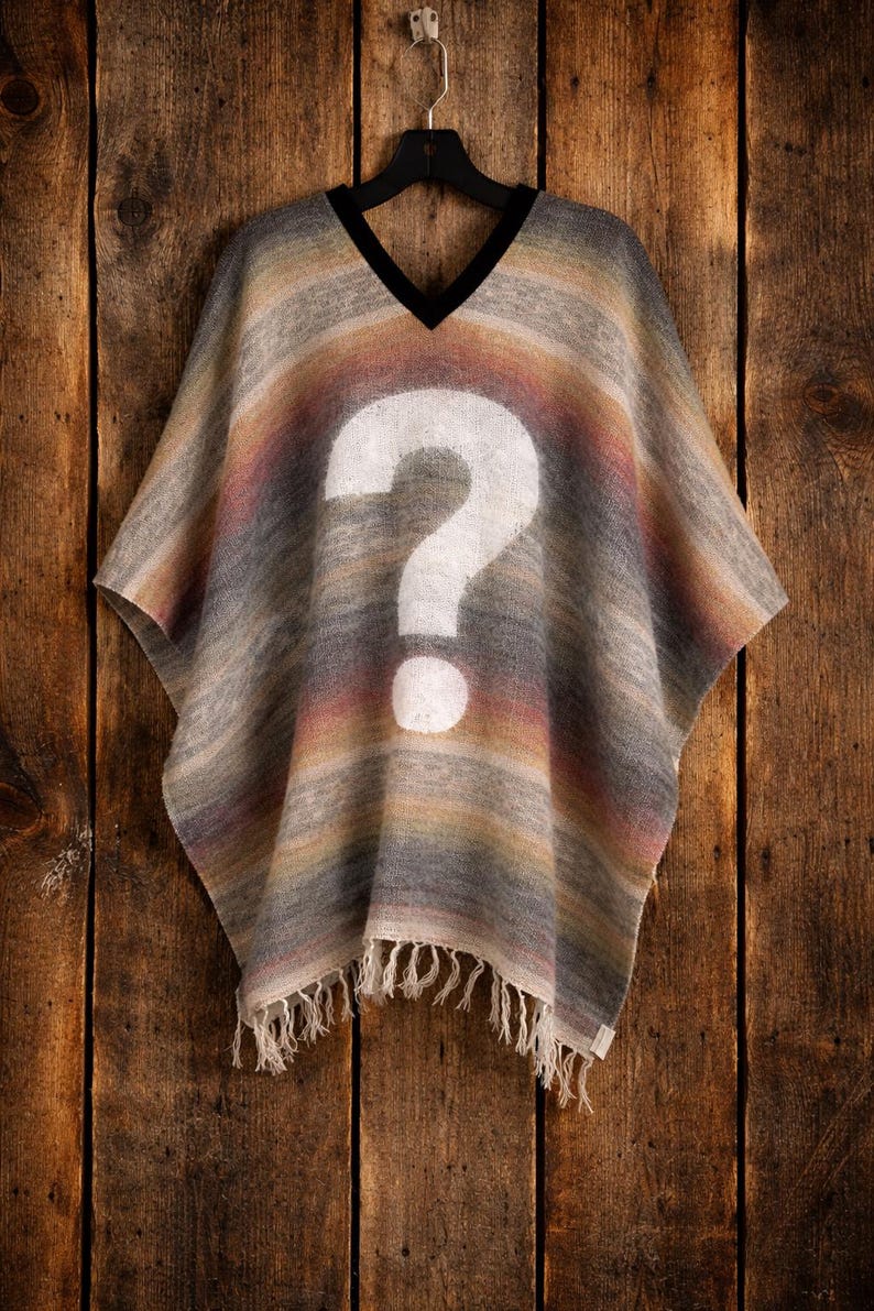 May include: A V-neck poncho featuring a horizontal gradient of muted earth tones including grey, tan, and soft pink. A large, white question mark is printed in the center of the garment. The hem is finished with a fringe of loose, off-white threads.