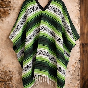 May include: A poncho featuring a woven pattern of horizontal stripes in shades of lime green, dark forest green, black, and white. The garment has a V-neckline with black trim and a fringed hemline at the bottom, hanging from a black hanger.