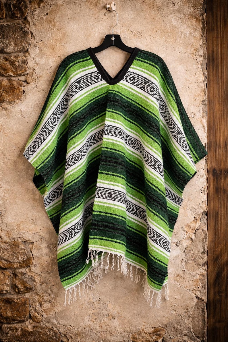 May include: A poncho featuring a woven pattern of horizontal stripes in shades of lime green, dark forest green, black, and white. The garment has a V-neckline with black trim and a fringed hemline at the bottom, hanging from a black hanger.