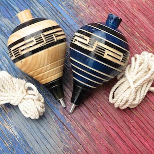 May include: Two wooden spinning tops, one natural wood and the other blue, with black stripes and a metal tip. Each top has a white rope. The tops are on a red and blue wooden surface.
