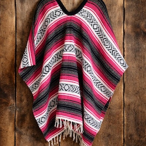 May include: A woven poncho featuring a vibrant pattern of horizontal stripes in hot pink, black, and white. The garment has a V-neckline with black trim and a fringe hem at the bottom. The fabric displays a repeating geometric diamond motif.