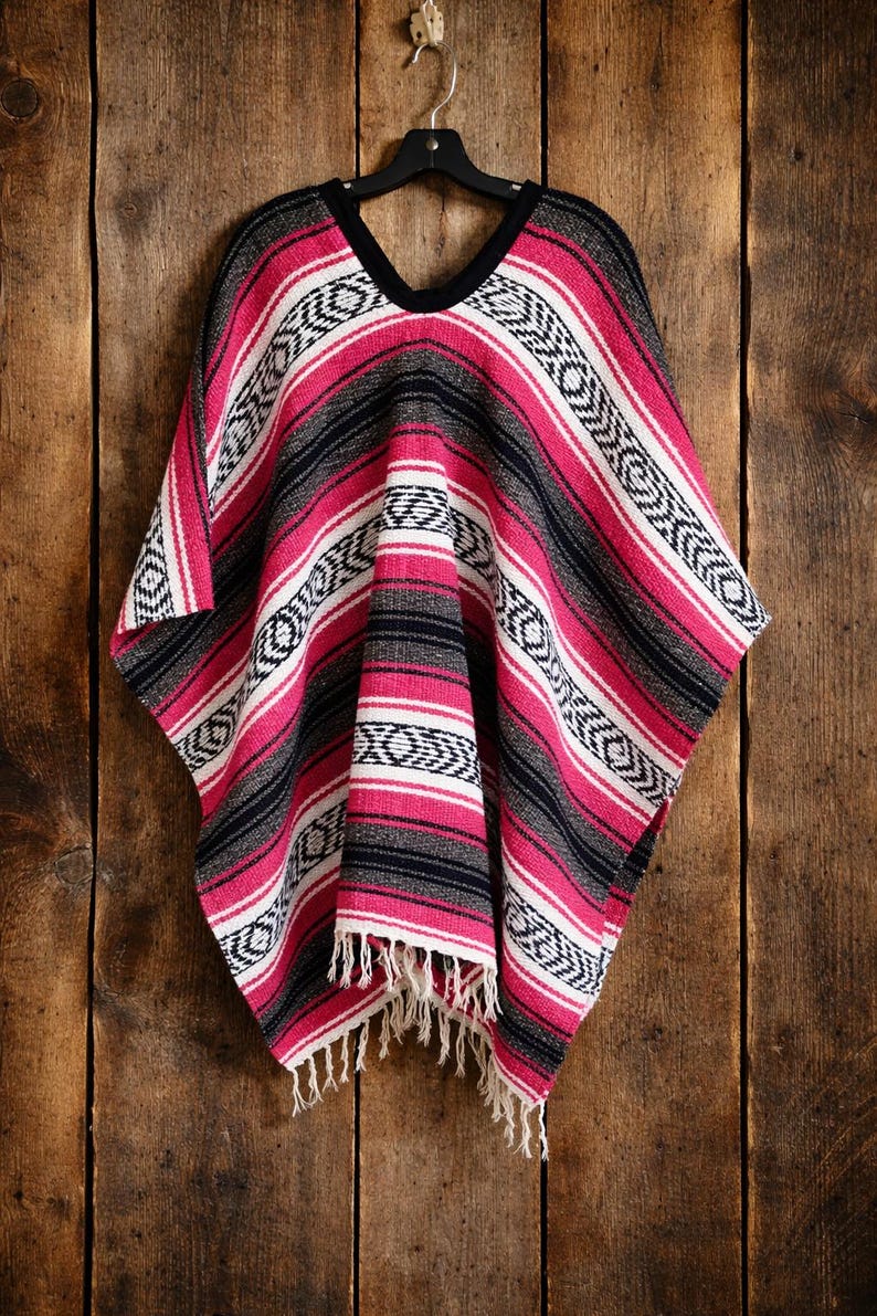 May include: A woven poncho featuring a vibrant pattern of horizontal stripes in hot pink, black, and white. The garment has a V-neckline with black trim and a fringe hem at the bottom. The fabric displays a repeating geometric diamond motif.