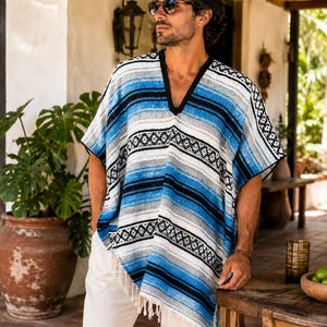 Könnte beinhalten: Ein V-Ausschnitt-Poncho mit einem Webmuster in Blau-, Weiß- und Schwarztönen. Das Kleidungsstück hat einen schwarzen Besatz am Ausschnitt und Fransen am unteren Saum, was durch die horizontalen Streifen eine strukturierte Optik erzeugt.