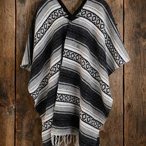May include: A woven poncho featuring a black, gray, and white striped pattern with a repeating geometric design. The garment has a V-neckline with black trim and a fringe hem at the bottom, hanging from a black plastic hanger.