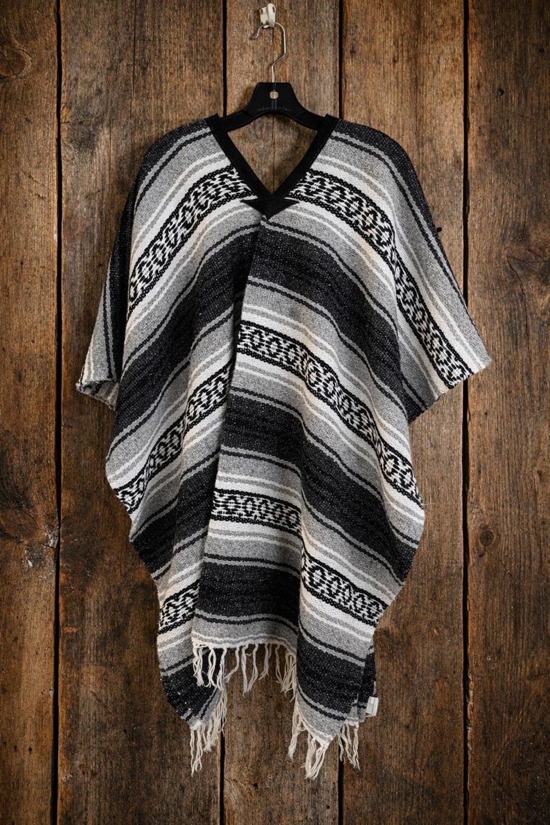 May include: A woven poncho featuring a black, gray, and white striped pattern with a repeating geometric design. The garment has a V-neckline with black trim and a fringe hem at the bottom, hanging from a black plastic hanger.
