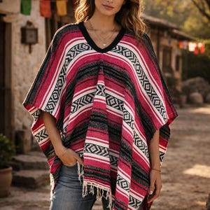 May include: A woven poncho featuring a vibrant pattern of horizontal stripes in shades of hot pink, black, and white. The garment has a V-neckline with a solid black trim, short sleeves, and a fringed hemline, paired with blue denim jeans.