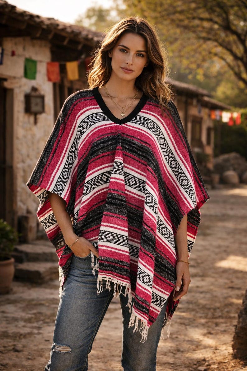May include: A woven poncho featuring a vibrant pattern of horizontal stripes in shades of hot pink, black, and white. The garment has a V-neckline with a solid black trim, short sleeves, and a fringed hemline, paired with blue denim jeans.