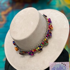 Puede incluir: Un sombrero de ala ancha de color beige claro con un adorno floral colorido. El adorno presenta una cadena de pequeñas flores y cuentas de colores. El sombrero está hecho de un material suave, similar al ante.