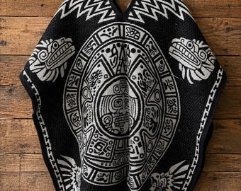 May include: A black and white poncho featuring a central circular Aztec calendar design, surrounded by stylized animal motifs and geometric patterns. The garment has a V-neckline, a black trim along the edges, and white fringe along the bottom hem.