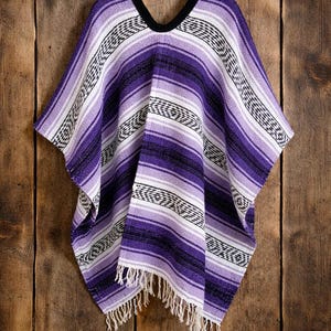 May include: A woven poncho featuring horizontal stripes in shades of purple, lavender, white, and black. The garment has a V-neckline with a black trim and a fringe hem at the bottom. The fabric displays a repeating geometric pattern throughout.