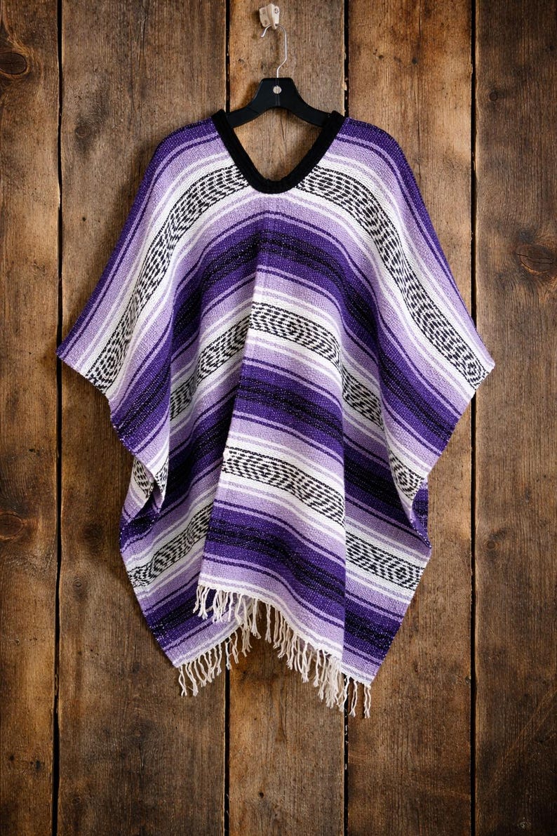 May include: A woven poncho featuring horizontal stripes in shades of purple, lavender, white, and black. The garment has a V-neckline with a black trim and a fringe hem at the bottom. The fabric displays a repeating geometric pattern throughout.