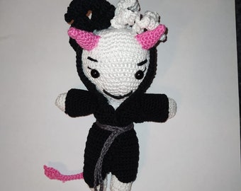 Peut inclure: Un jouet en peluche crocheté avec un corps blanc, des cheveux noirs et blancs, des cornes roses et une robe noire avec une ceinture grise. Le jouet a une queue rose.