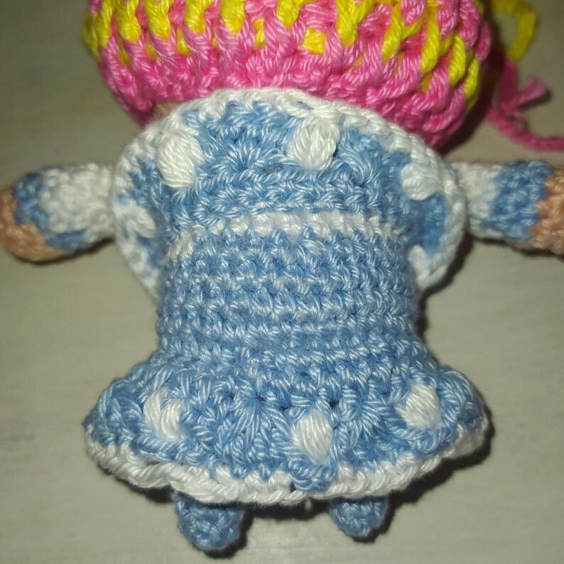 Pode incluir: Uma boneca de croch&ecirc; vestindo um vestido azul com bolinhas brancas e um chap&eacute;u rosa e amarelo.