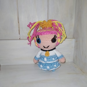May include: A crocheted doll with pink and yellow hair, blue eyes, and a blue and white dress. The doll is wearing a yellow necklace and has a mischievous smile.