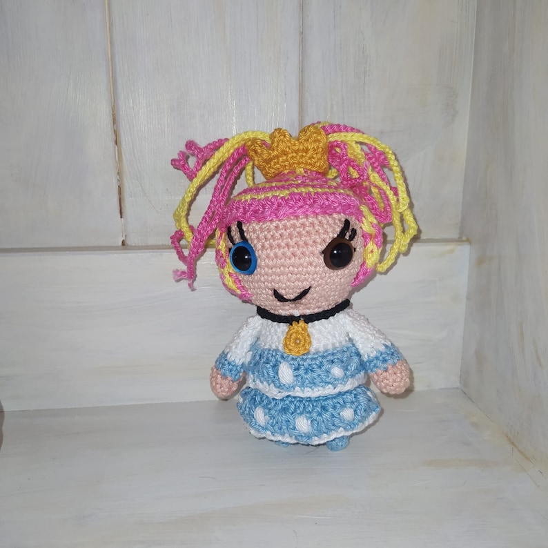 Pode incluir: Uma boneca de croch&ecirc; com cabelo rosa e amarelo, olhos azuis e um vestido azul e branco. A boneca est&aacute; usando um colar amarelo e tem um sorriso travesso.