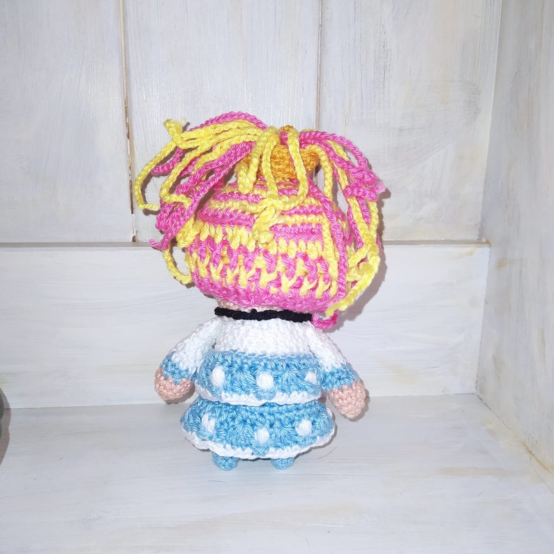 Pode incluir: Uma boneca de croch&ecirc; com cabelo rosa e amarelo, vestindo um su&eacute;ter branco e um vestido azul com bolinhas brancas.