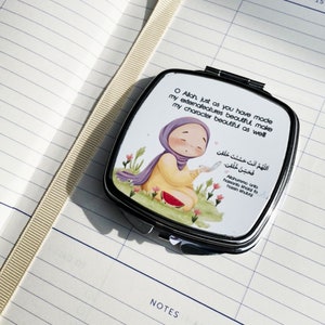 May include: A silver compact mirror with a cartoon illustration of a person in a hijab, kneeling in a field of flowers. The mirror has text in English and Arabic. The mirror is open and resting on a lined notebook.